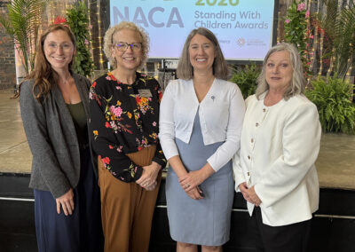 For women of various ages are dressed professionally and pose in front of a projector screen that reads NACA 2026.