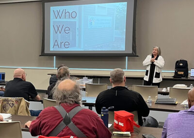 Rows of adults sit in a classroom type setting with a presentation showing on a projection. CEO Brenda Porter is seen at the front presenting a slide about "Who We Are".