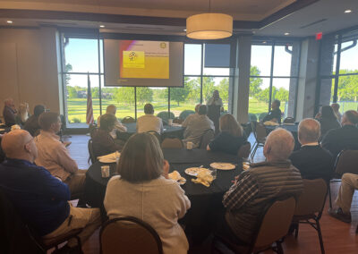 A large gathering of adults sit at various round tables in a large room with a wall of windows. A presentation can be seen where a CACCMO staff member is sharing the nonprofit's story and mission.